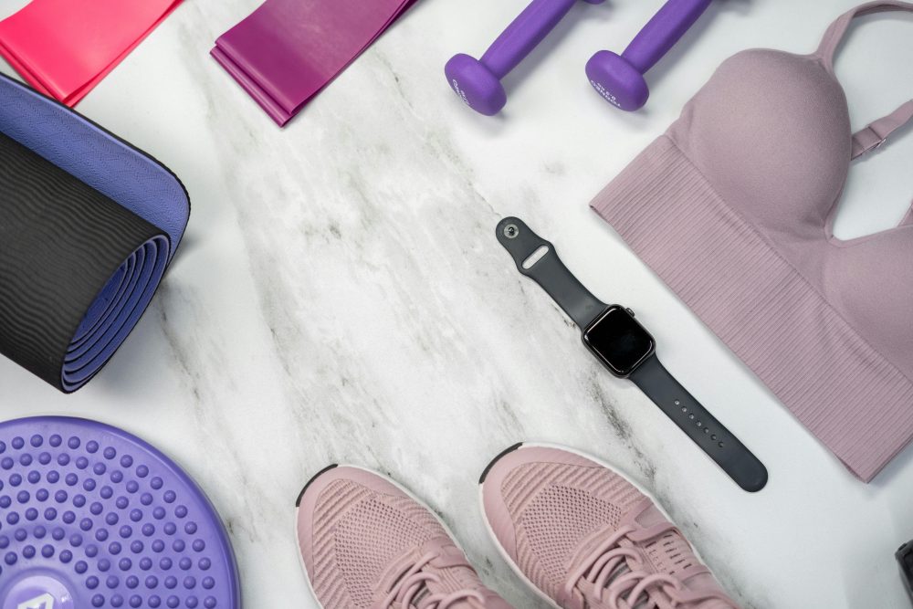 Home-gym starter kit: yoga mat, resistance bands, light dumbbells, sports bra, smartwatch, sneakers on marble backdrop