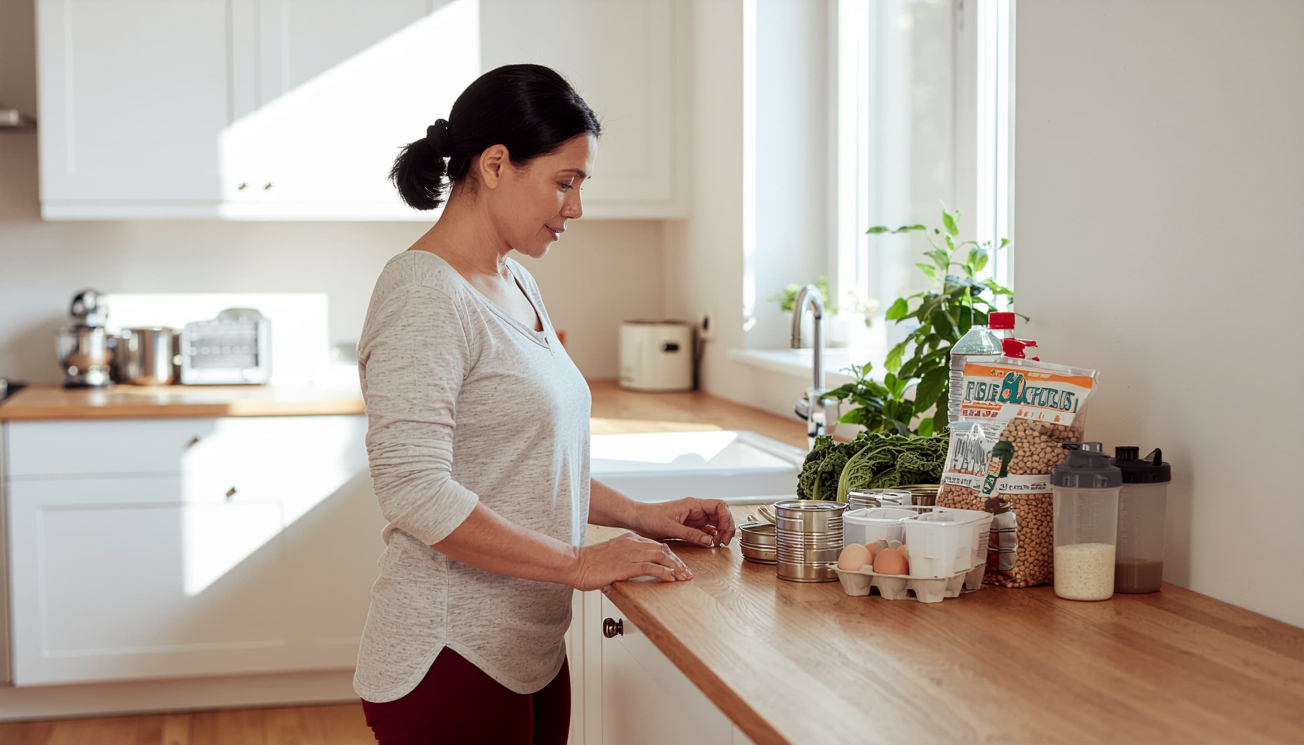 Woman in her late 40s standing at a kitchen counter with affordable high-protein groceries — canned tuna, lentils, Greek yogurt, eggs, and a protein shake