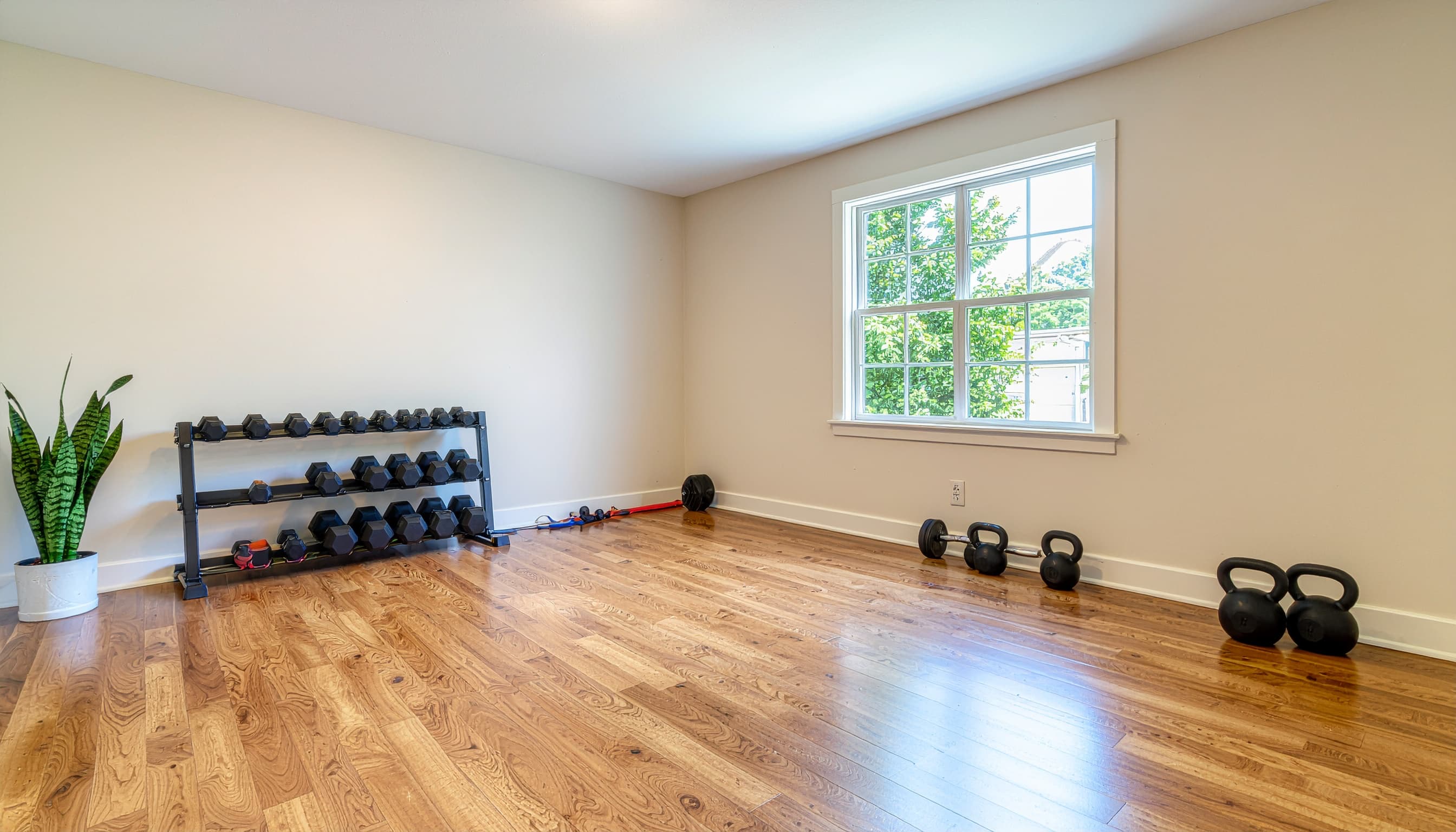 Bright converted home gym with dumbbell rack, hardwood floor, and natural light from a large window